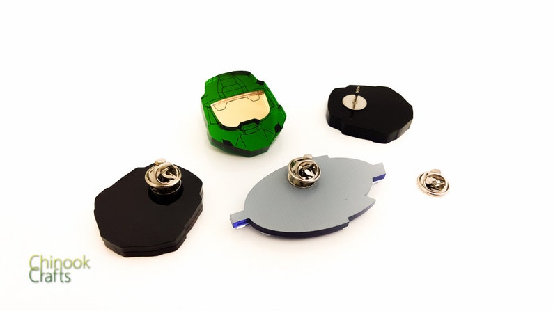 Pode incluir: Uma cole&ccedil;&atilde;o de alfinetes de acr&iacute;lico em v&aacute;rias formas e cores. Um alfinete &eacute; um capacete verde e dourado, outros s&atilde;o pretos, cinzentos e azuis. Cada alfinete tem um verso prateado. O logotipo Chinook Crafts est&aacute; no canto inferior esquerdo.