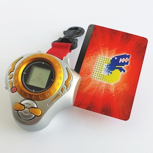 Peut inclure: Un appareil numérique pour animaux de compagnie argenté et doré avec une sangle rouge et une carte bleue et jaune avec un motif de circuit imprimé et un graphique de dinosaure bleu. La carte indique "Digi-Monster".