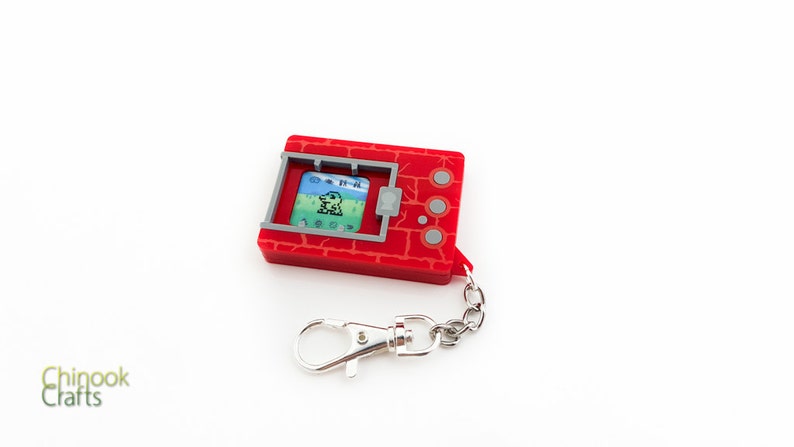 K&ouml;nnte beinhalten: Roter und grauer digitaler Haustier-Schl&uuml;sselanh&auml;nger mit einem kleinen Bildschirm, auf dem eine pixelartige gr&uuml;ne Kreatur zu sehen ist. Der Schl&uuml;sselanh&auml;nger hat eine silberne Kette und einen silbernen Karabiner.
