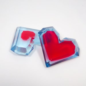 Könnte beinhalten: Zwei herzförmige Harz-Ornamente, eines ist blau mit einem roten Herzen im Inneren, das andere ist rot mit einem blauen Umriss.