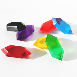 Könnte beinhalten: Sechs bunte geometrische Formen aus Kunststoff, die an Edelsteine erinnern, in verschiedenen Farben, darunter Rot, Blau, Grün, Gelb, Lila und Schwarz.