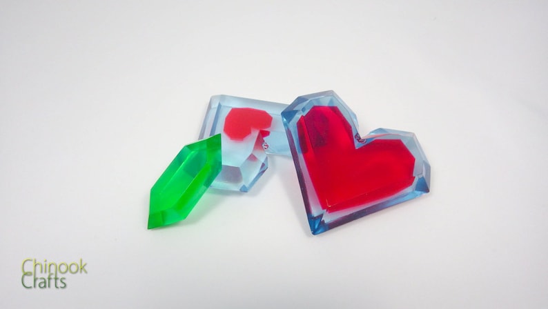 Pu&ograve; includere: Tre cuori in resina, uno verde e due rossi, con uno strato esterno blu trasparente. I cuori sono su uno sfondo bianco.