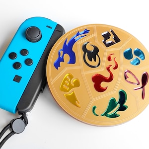May include: A gold round disc with a variety of colorful symbols on it. The disc is next to a blue Nintendo Switch controller.