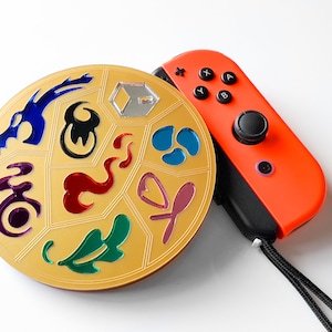 Puede incluir: Un disco redondo de color dorado con varios símbolos coloridos, incluyendo un dragón, llamas y una hoja. El disco está al lado de un mando Nintendo Switch Joy-Con rojo y negro.