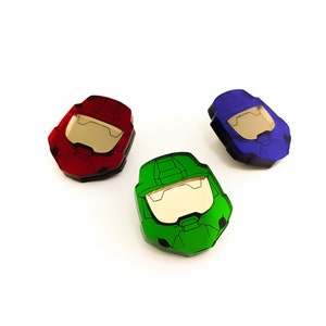 Pode incluir: Tr&ecirc;s objetos coloridos em forma de capacete, em vermelho, verde e azul, s&atilde;o exibidos em um fundo branco. Cada capacete tem uma viseira dourada. Os objetos parecem ser feitos &agrave; m&atilde;o, possivelmente de papel ou cart&atilde;o, e est&atilde;o dispostos em grupo.