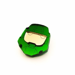 Pode incluir: Um objeto verde em forma de capacete angular com uma viseira dourada, que lembra um personagem de videogame. O objeto est&aacute; sobre um fundo branco, com o texto "Chinook Crafts" no canto inferior esquerdo.