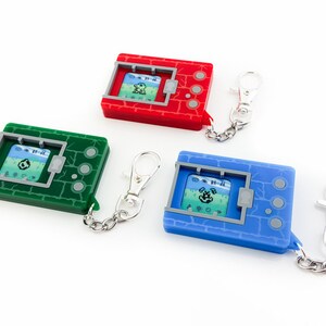 K&ouml;nnte beinhalten: Drei digitale Haustier-Schl&uuml;sselanh&auml;nger in Rot, Gr&uuml;n und Blau. Jedes Spielzeug hat einen kleinen Bildschirm und Kn&ouml;pfe. Die Spielzeuge sind an silbernen Ketten befestigt.