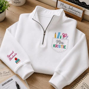 May include: A white quarter-zip sweatshirt featuring an embroidered pocket design with school supplies and the name Mrs. KRISTIE. The sleeve displays the text Special Students above an illustration of a red apple resting on a stack of books.
