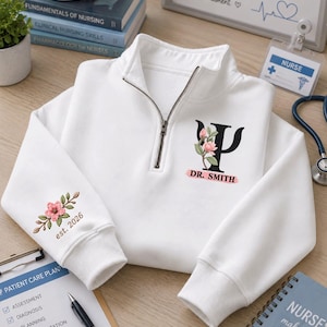 May include: A white quarter-zip sweatshirt featuring a black Greek psi symbol adorned with a pink flower graphic and the text DR. SMITH on the chest. The right sleeve displays a pink floral embroidery with the text est. 2026 in brown lettering.