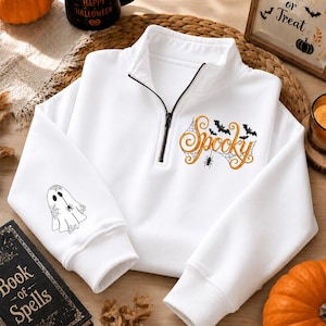 Puede incluir: Una sudadera blanca con cremallera corta y un bordado en color naranja con la palabra Spooky, decorada con murciélagos negros, una araña y una telaraña. La manga izquierda muestra un fantasma envuelto en una telaraña con una pequeña araña.