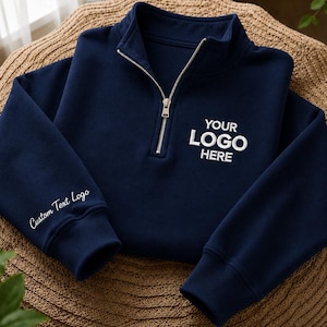 Könnte beinhalten: Ein marineblaues Sweatshirt mit Viertelreißverschluss und silberfarbenem Metallreißverschluss am Kragen. Auf der Brust befindet sich der weiß gestickte Text YOUR LOGO HERE, am rechten Ärmelbündchen der weiß gestickte Text Custom Text Logo.