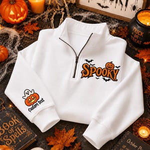 May include: A white quarter-zip sweatshirt featuring the orange text "SPOOKY" with a pumpkin and bats on the chest. The right sleeve displays a graphic of a ghost sitting on a pumpkin with the words "Custom text" printed underneath in black.