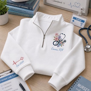 May include: A white quarter-zip sweatshirt featuring an embroidered stethoscope intertwined with colorful flowers on the chest, with the text "Emma, RN" below. The sleeve displays a red medical cross, a heartbeat line, and the text "Est. 2026".