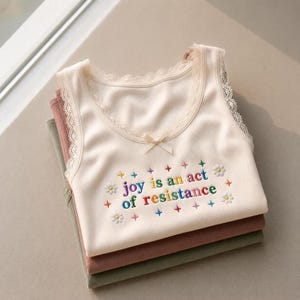Puede incluir: Una camiseta de tirantes acanalada de color crema con ribete de encaje a lo largo del escote y las sisas, con un pequeño lazo central y texto bordado que dice joy is an act of resistance, rodeado de flores y estrellas de colores.