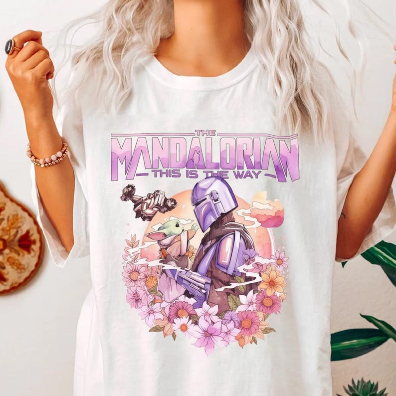 May include: White t-shirt featuring a graphic design from The Mandalorian. The design includes the text "The Mandalorian - This is the Way" in purple and white, along with illustrations of the Mandalorian, Grogu, and a ship, surrounded by flowers.