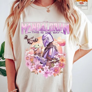 May include: Cream-colored t-shirt featuring a graphic of The Mandalorian and Grogu surrounded by flowers. The design includes the text "THE MANDALORIAN - THIS IS THE WAY" in purple and white. The artwork has a vintage, floral aesthetic.