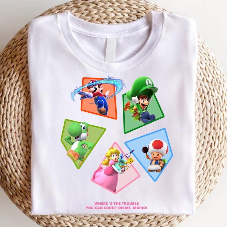 May include: White t-shirt featuring colorful cartoon characters Mario, Luigi, Yoshi, Peach, and Toad in geometric shapes. The text "WHERE'S THE TROUBLE YOU CAN COUNT ON ME, MARIO!" is printed at the bottom. The shirt is folded on a woven surface.