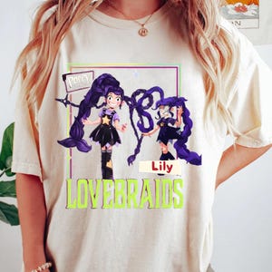 Puede incluir: Camiseta color crema con un gráfico de dos chicas de dibujos animados con largas trenzas moradas. Los textos "Poppy" y "Lily" están encima de las chicas, y la palabra "LOVEBRAIDS" está debajo en letras verdes.