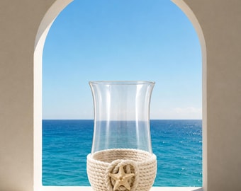 May include: A clear glass hurricane candle holder featuring a base wrapped in thick, cream-colored nautical rope. A small, textured beige starfish is attached to the center of the rope wrapping, providing a coastal accent to the cylindrical glass vessel.