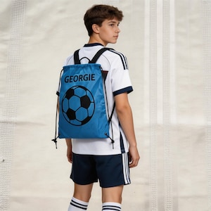 May include: A blue drawstring backpack with a black soccer ball graphic and the name "GEORGIE" in black. The backpack is worn by a person wearing a white and navy blue soccer uniform.