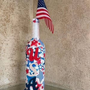 May include: A decorative bottle with a patriotic theme. The bottle is adorned with red, white, and blue stars and stripes, with the letters "USA" prominently displayed. A small American flag is attached to the top, making it a festive item.