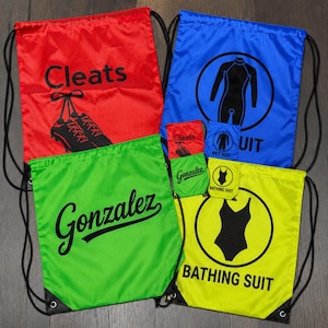 Op de afbeelding: Een verzameling van vier rugzakken met trekkoord in rood, blauw, groen en geel. De rode tas bevat de tekst Cleats en een afbeelding van schoenen. De blauwe tas toont een wetsuit-pictogram en tekst. De groene tas toont de naam Gonzalez. De gele tas toont een badpak-pictogram en tekst.