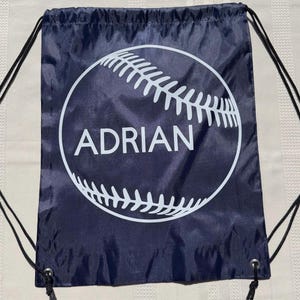 Peut inclure: Un sac à cordon bleu marine arborant un grand motif blanc représentant une balle de baseball avec le nom ADRIAN imprimé au centre. Le sac est doté de cordons noirs fixés à des œillets métalliques aux coins inférieurs pour la fermeture.