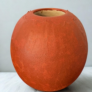 Puede incluir: Un jarrón redondo de cerámica naranja con una superficie texturizada. El jarrón tiene una abertura ancha y un interior de color más claro. El exterior es de color naranja mate sólido. El jarrón es un objeto decorativo.