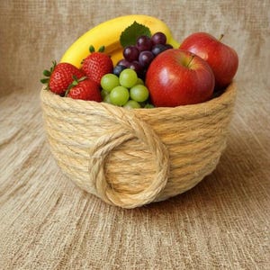 May include: A round bowl crafted from coiled natural tan jute rope filled with fresh fruit including a yellow banana, two bright red strawberries, a cluster of small green grapes, a bunch of dark purple grapes, and two shiny red apples.