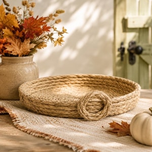 May include: A round, shallow tray constructed from coiled natural tan jute rope, featuring a single circular rope handle attached to the side. The tray has a textured, woven appearance with a consistent thickness throughout its circular frame.