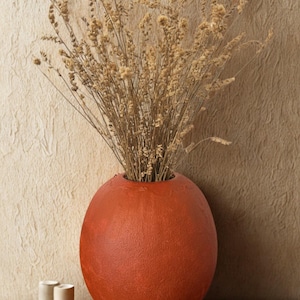 Könnte beinhalten: Eine große, runde Vase in Terrakotta-Farbe, gefüllt mit getrockneten, beigefarbenen Ziergräsern. Die Vase steht auf einer Holzoberfläche mit zwei kleinen, zylindrischen, cremefarbenen Kerzen. Der Hintergrund ist eine strukturierte, beige Wand.