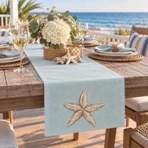 May include: A light blue table runner featuring a large, embroidered starfish design in beige and white tones. The runner is placed on a wooden table set with white plates, blue napkins, wine glasses, and a centerpiece of white flowers.