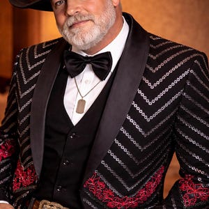 Könnte beinhalten: Ein Mann trägt einen schwarzen Cowboyhut, einen schwarzen Paillettenblazer mit roter Stickerei und eine schwarze Fliege. Der Blazer hat ein Zickzackmuster. Er trägt auch eine goldene Gürtelschnalle.