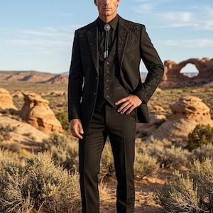 Puede incluir: Un traje de tres piezas negro que incluye una chaqueta con bordados tonales en las solapas y puños, un chaleco a juego, una camisa negra abotonada, una corbata de bolo con un broche plateado y botas de cuero negro con punta afilada.