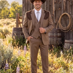 Könnte beinhalten: Ein dreiteiliger brauner Anzug mit dekorativen Nähten auf der Jacke, kombiniert mit einem weißen Hemd und einer Bolo-Krawatte. Das Ensemble wird durch einen braunen Cowboyhut und Stiefel ergänzt. Der Mann steht in einem Feld mit einer Holzkonstruktion im Hintergrund.
