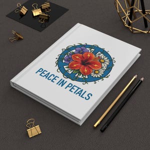 Könnte beinhalten: Weißes Notizbuch mit dem Text "PEACE IN PETALS" und einem floralen Peace-Zeichen-Design. Das Design zeigt eine rote Hibiskusblüte, lila und weiße Blumen und grüne Ranken. Goldene und schwarze Bleistifte und goldene Büroklammern sind ebenfalls sichtbar.