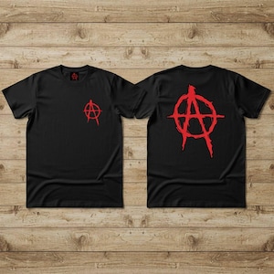 May include: Black t-shirts with a red anarchy symbol. One shirt displays the symbol on the front left chest, and the other has a larger version on the back. The shirts are displayed on a wooden surface.