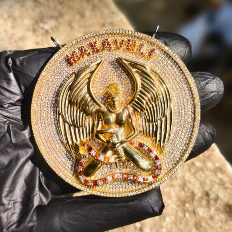May include: A gold-colored pendant with the word "MAKAVELI" in raised letters. The pendant features a winged figure with a serpent design and is encrusted with clear and red gemstones. The pendant is round and has a loop at the top.