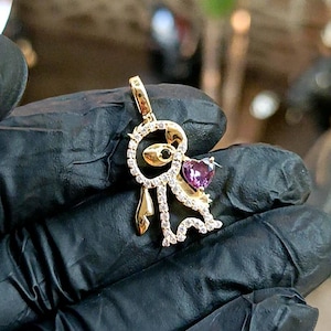 May include: A gold-colored pendant featuring a stylized figure with a heart-shaped purple gemstone. The figure is adorned with small, clear gemstones and a black gemstone eye. The pendant has a loop for a chain.