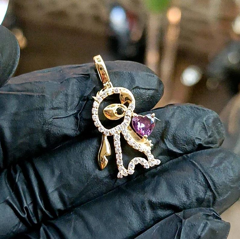 May include: A gold-colored pendant featuring a stylized figure with a heart-shaped purple gemstone. The figure is adorned with small, clear gemstones and a black gemstone eye. The pendant has a loop for a chain.