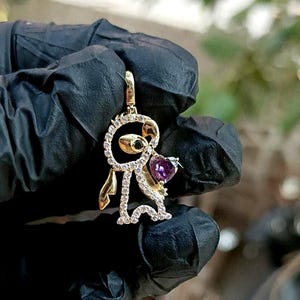 May include: A gold-colored pendant in the shape of a cartoon character, featuring a purple heart-shaped gemstone. The pendant is adorned with small, clear gemstones and has a loop for a chain. The character has a black eye and gold-colored accents.