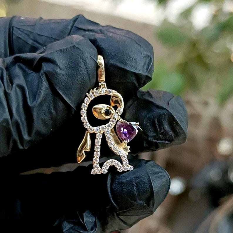 May include: A gold-colored pendant in the shape of a cartoon character, featuring a purple heart-shaped gemstone. The pendant is adorned with small, clear gemstones and has a loop for a chain. The character has a black eye and gold-colored accents.