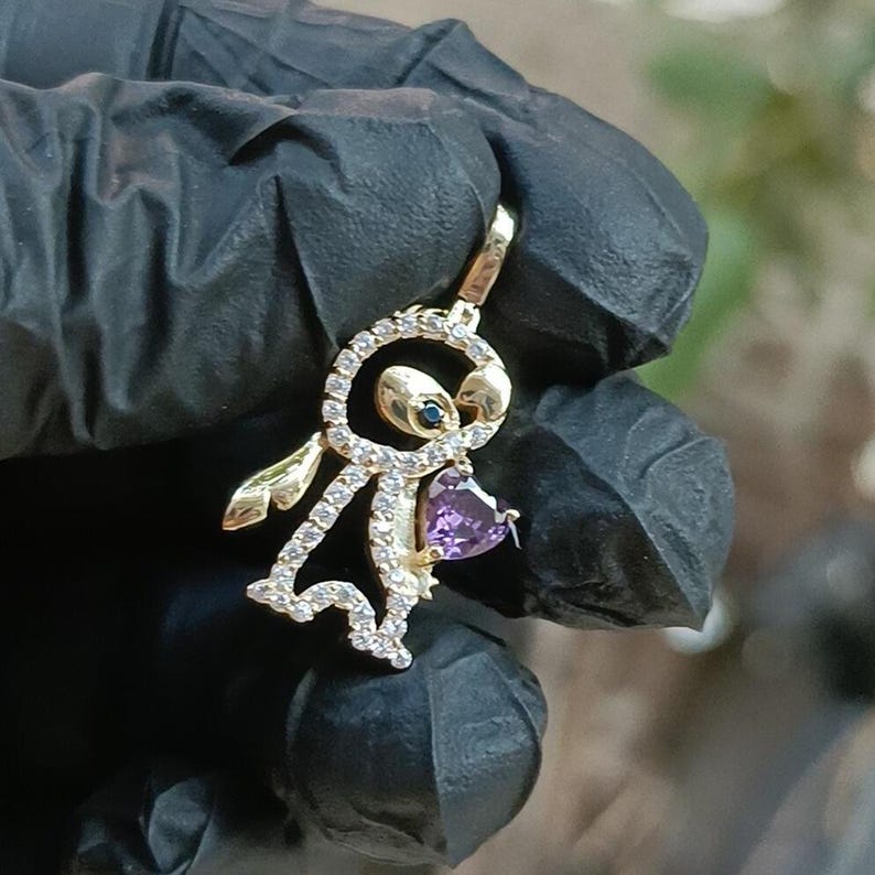 May include: A gold-colored pendant in the shape of a cartoon character, encrusted with clear crystals. The pendant features a purple heart-shaped gem and a small blue gem. The pendant is held against a black background.