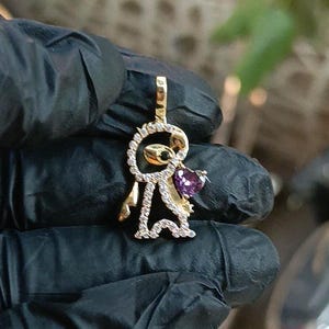 May include: A gold-colored pendant in the shape of a stylized figure, encrusted with small clear stones. The figure holds a heart-shaped purple gemstone. The pendant is held by a gloved hand.