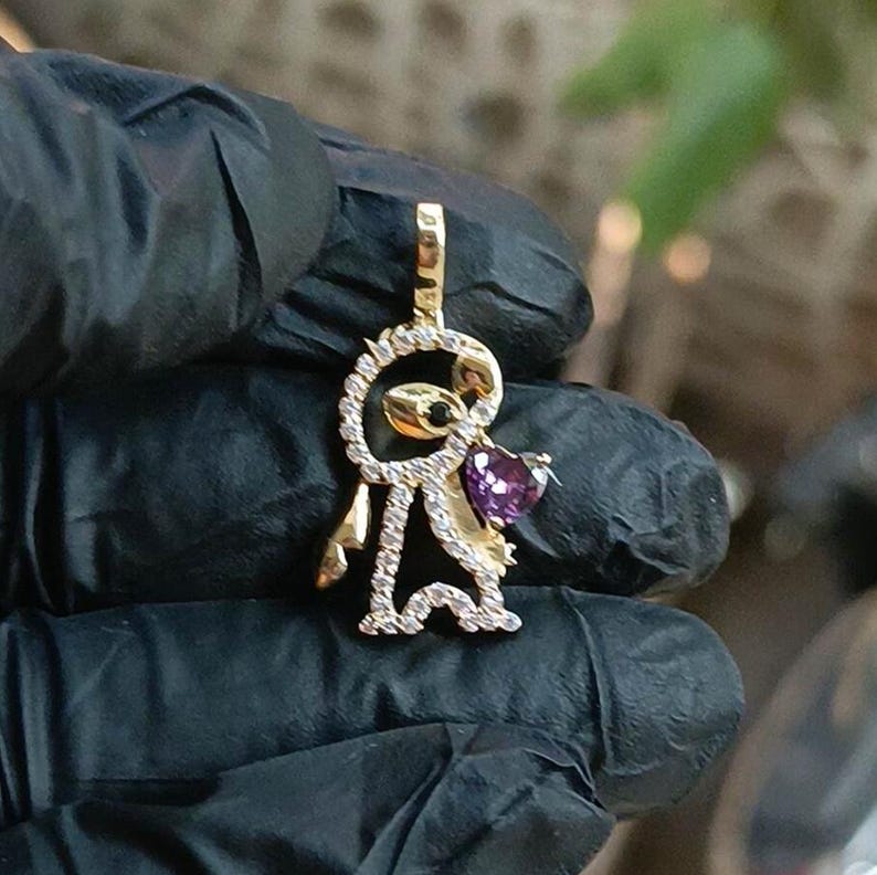 May include: A gold-colored pendant in the shape of a stylized figure, encrusted with small clear stones. The figure holds a heart-shaped purple gemstone. The pendant is held by a gloved hand.