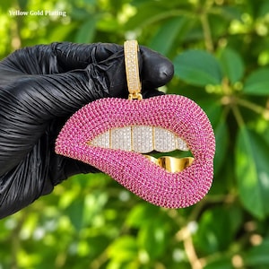Puede incluir: Un colgante chapado en oro amarillo con forma de labios, cubierto de pequeñas piedras rosas. Los dientes superiores están incrustados con piedras blancas transparentes, mientras que los inferiores son de oro amarillo liso. Una reasa rectangular superior tiene piedras.