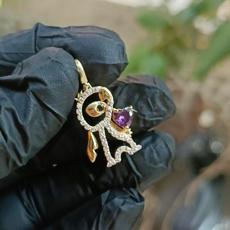 May include: A gold-colored pendant featuring a stylized dog design. The pendant has a heart-shaped purple gemstone and a small green gemstone. The dog's body is outlined with small, clear gemstones. The pendant is suspended from a small loop.