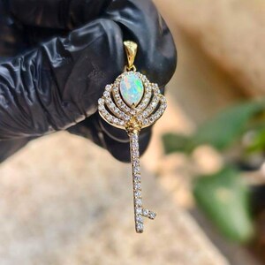 Puede incluir: Colgante de llave dorada con una gran piedra preciosa de ópalo ovalada. La corona de la llave está adornada con pequeñas piedras claras y brillantes. El vástago y el paletón de la llave también están adornados con piedras claras.