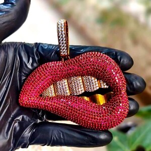 May include: A pendant shaped like a pair of lips, encrusted with red gemstones. The teeth are gold-colored, with some teeth featuring white gemstones. The bail is also covered in white gemstones. The pendant is held by a gloved hand.