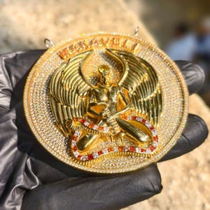 May include: A gold pendant featuring a winged figure with intricate detailing. The pendant is encrusted with numerous small, sparkling stones and has a red and white accent. The word "RENAISSANCE" is visible on the top of the pendant.